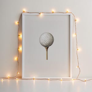 May include: A framed print featuring a golf ball on a tee. The golf ball is white with a dimpled surface, and the tee is a light brown color. The frame is white, and the image is decorated with a string of small, warm-toned lights.
