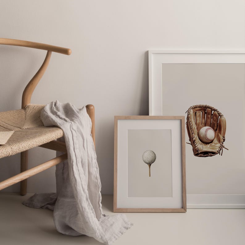 May include: A framed print of a golf ball on a tee, a baseball glove with a baseball, and a wooden chair with a light-colored throw. The artwork has a neutral color palette and a minimalist aesthetic.