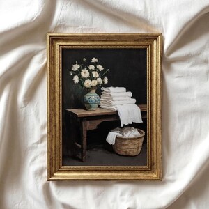 May include: A framed painting depicts a still life scene. A vase of white flowers sits on a wooden bench next to a stack of white towels. A wicker basket holds more white linens. The painting is in a gold frame.