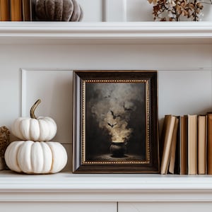 May include: A framed artwork with a Halloween theme, depicting a cauldron with smoke and bats. The frame is dark wood with a gold inner edge. Two white pumpkins and a stack of books are on the shelf. The color palette is dark with gold accents.