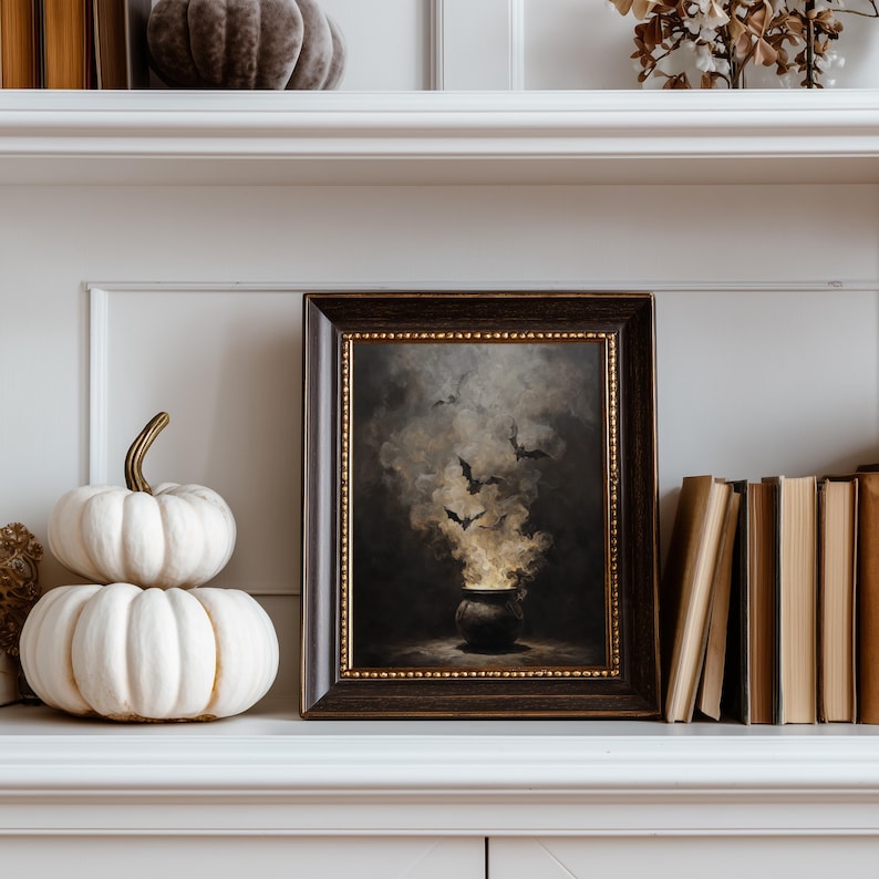 May include: A framed artwork with a Halloween theme, depicting a cauldron with smoke and bats. The frame is dark wood with a gold inner edge. Two white pumpkins and a stack of books are on the shelf. The color palette is dark with gold accents.
