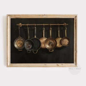 Puede incluir: Una impresi&oacute;n art&iacute;stica enmarcada que muestra siete sartenes de metal variadas colgadas de una barra horizontal sobre un fondo negro s&oacute;lido. Las sartenes var&iacute;an en tama&ntilde;o y acabado met&aacute;lico, desde lat&oacute;n pulido hasta hierro oscuro, todas suspendidas por ganchos.