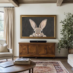 May include: A framed painting of a brown rabbit peeking out from behind a dark gray background. The rabbit has long ears and a white nose. The frame is gold.