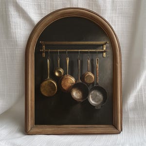 May include: A framed artwork featuring a collection of antique copper and brass cooking pots and pans hanging from a brass rod. The pots vary in size and shape, with a dark background and a wooden frame.