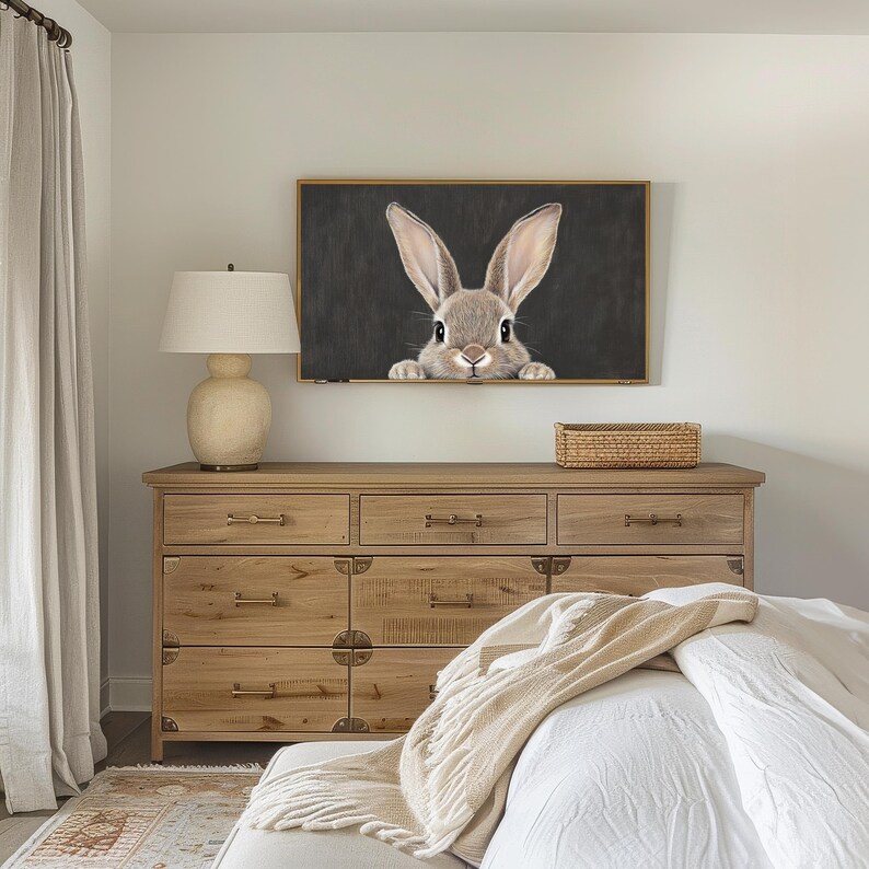 May include: A wooden dresser with six drawers and brass hardware. A framed print of a rabbit peeking out from behind a dark background hangs above the dresser. A white bed with a beige throw blanket is in the foreground.