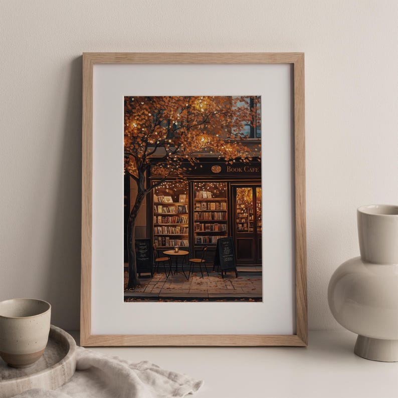 May include: A framed print of a cozy book cafe scene. The cafe has a dark brown facade with the words "Book Cafe" above the entrance. The interior is lit with warm lights, and a tree with orange leaves frames the scene.