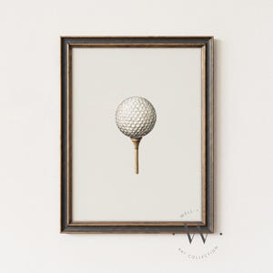 May include: A framed print featuring a golf ball on a tee. The golf ball is white with a dimpled surface, and the tee is a light brown color. The frame is dark with gold accents. The print is from the Well Art Collection.