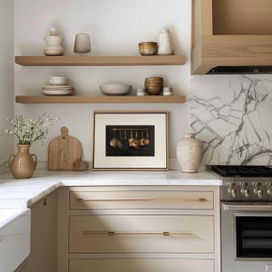 Puede incluir: Una escena de cocina con dos estantes flotantes de madera que contienen varios cuencos de cer&aacute;mica, jarrones y cristaler&iacute;a. Debajo, una impresi&oacute;n enmarcada de sartenes de cobre cuelga sobre una encimera blanca junto a una tabla de madera.