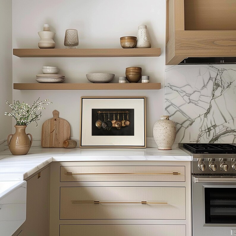 Puede incluir: Una escena de cocina con dos estantes flotantes de madera que contienen varios cuencos de cer&aacute;mica, jarrones y cristaler&iacute;a. Debajo, una impresi&oacute;n enmarcada de sartenes de cobre cuelga sobre una encimera blanca junto a una tabla de madera.