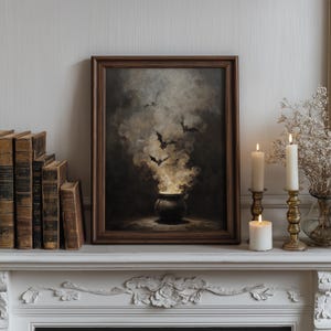 May include: A framed artwork with a Halloween theme, showing a cauldron with smoke and bats. The dark brown frame surrounds the image, set on a white mantelpiece. Lit candles in brass holders and antique books complete the scene.