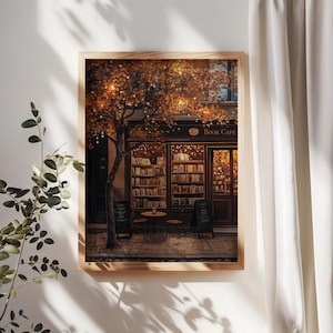 May include: A framed print of a cozy book cafe scene. The cafe, with the text "Book Cafe" above the entrance, is framed by a tree with autumn-colored leaves and string lights. Two small tables and chairs are set outside. The print is in a wooden frame.