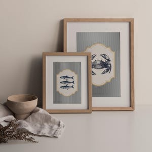 May include: Two framed nautical art prints. One print features three fish, and the other a crab, both in navy blue on a striped background. The frames are light brown wood, and the prints are displayed on a white surface.