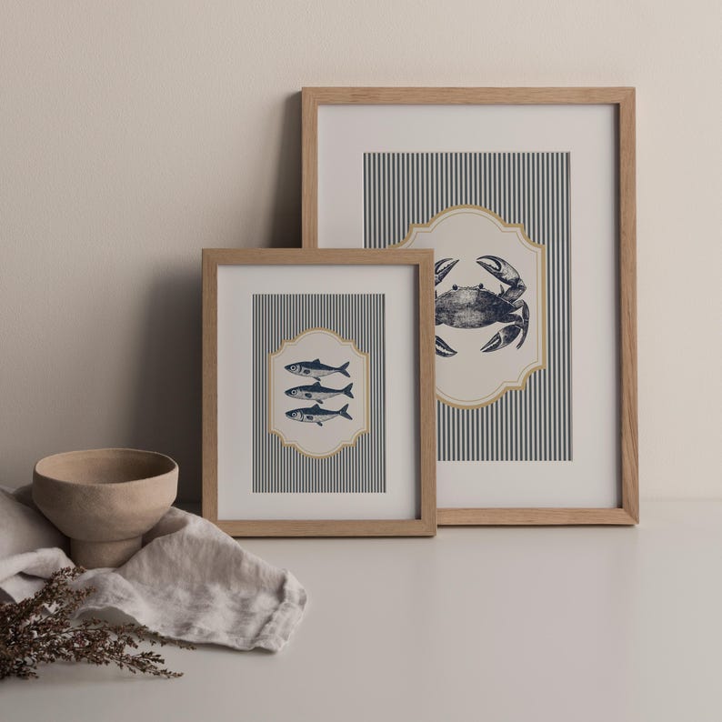 May include: Two framed nautical art prints. One print features three fish, and the other a crab, both in navy blue on a striped background. The frames are light brown wood, and the prints are displayed on a white surface.