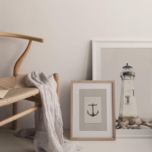 May include: A framed print with a black anchor illustration on a striped background. A second framed print shows a white lighthouse with a black top. A light-colored scarf drapes over a wooden chair.