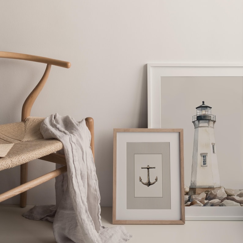 May include: A framed print with a black anchor illustration on a striped background. A second framed print shows a white lighthouse with a black top. A light-colored scarf drapes over a wooden chair.
