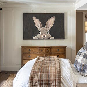 May include: A framed print of a brown rabbit peeking out from behind a dark gray background. The print is hanging on a white wall above a wooden dresser with a white bedspread and a brown plaid throw blanket.