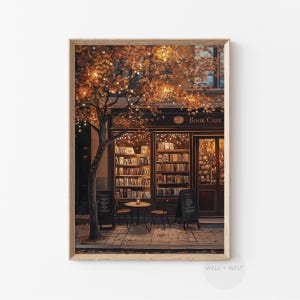 May include: A framed print of a cozy book cafe scene. The storefront features a dark facade with the words "Book Cafe" above the entrance. A tree with orange leaves and string lights frames the scene. Two small black signs and a table with chairs are in front.