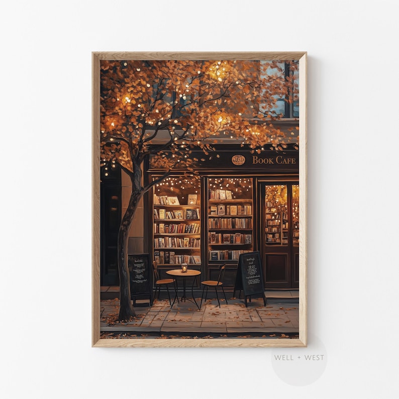 May include: A framed print of a cozy book cafe scene. The storefront features a dark facade with the words "Book Cafe" above the entrance. A tree with orange leaves and string lights frames the scene. Two small black signs and a table with chairs are in front.
