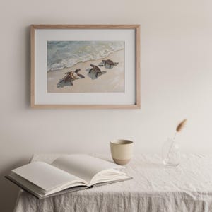 Op de afbeelding: Een ingelijst aquarel schilderij van drie zeeschildpadden die op een zandstrand lopen met oceaan golven op de achtergrond.