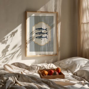 May include: A framed art print showing three blue fish on a cream background, set against navy and white vertical stripes. The print is in a light wood frame, creating a nautical theme. The print is in a light wood frame.