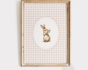 May include: Framed art print featuring a watercolor illustration of a brown bunny holding pink flowers. The bunny is centered on an oval frame with a dotted border, set against a pink and white gingham background. The frame is a light wood color.
