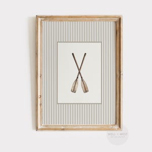 Puede incluir: Lámina enmarcada con dos remos de madera cruzados. Los remos están representados en acuarela sobre un fondo blanco, enmarcados por un borde gris y un patrón de rayas. El marco es de color marrón claro.