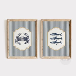 May include: Two framed nautical art prints. One features a detailed crab illustration, the other showcases three fish. Both are set against a blue and white striped background, with a cream-coloured, ornate border and a wooden frame.