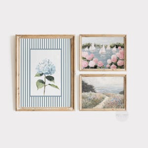May include: Set of three framed art prints. The prints feature watercolor paintings of a blue hydrangea, sailboats on a lake surrounded by pink flowers, and a coastal path. The frames are a light wood color.