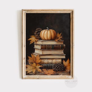May include: A framed autumn-themed print featuring a stack of old books, a pumpkin, pine cones, and fall leaves. The artwork has a warm color palette of browns, oranges, and greens, evoking a cozy, seasonal atmosphere. The frame is a light, neutral color.