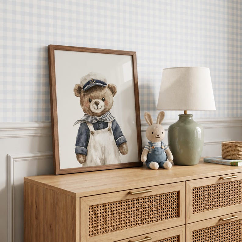 May include: A framed wall art print featuring a watercolor illustration of a brown teddy bear dressed as a sailor in a white cap, blue shirt, and white overalls. The bear has a friendly expression and is positioned next to a small stuffed rabbit.