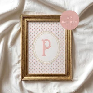 May include: A rectangular gold-toned frame containing a print with a repeating pattern of tiny pink flowers on a white background. Centered is a large pink letter P inside an oval decorative border. A pink circle overlay displays A-Z INCLUDED.