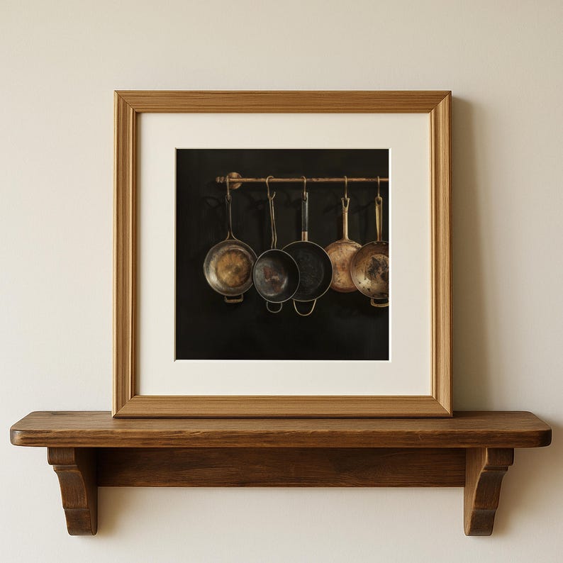 Puede incluir: Una obra de arte mural cuadrada enmarcada que muestra una fotograf&iacute;a de cinco sartenes met&aacute;licas colgadas de una barra horizontal sobre un fondo oscuro. El marco es de madera clara con un paspart&uacute; blanco, sobre un estante de madera oscura.