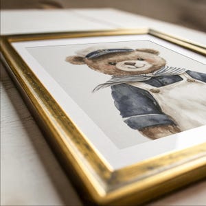 May include: A framed watercolor illustration of a brown teddy bear wearing a navy blue sailor shirt, a white sailor hat, and a white overall with buttons. The artwork is encased in a metallic gold frame with a white mat border, presented at an angle.