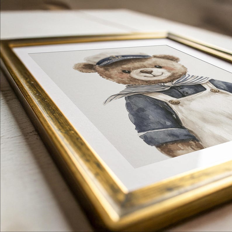 May include: A framed watercolor illustration of a brown teddy bear wearing a navy blue sailor shirt, a white sailor hat, and a white overall with buttons. The artwork is encased in a metallic gold frame with a white mat border, presented at an angle.