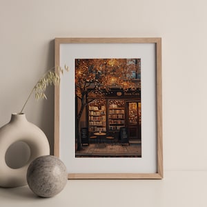 May include: A framed print of a cozy book cafe scene. The image features a storefront with the words "Book Cafe" above the entrance, bookshelves, a small table, and chairs. The scene is illuminated by warm lighting and surrounded by a tree with autumn leaves.