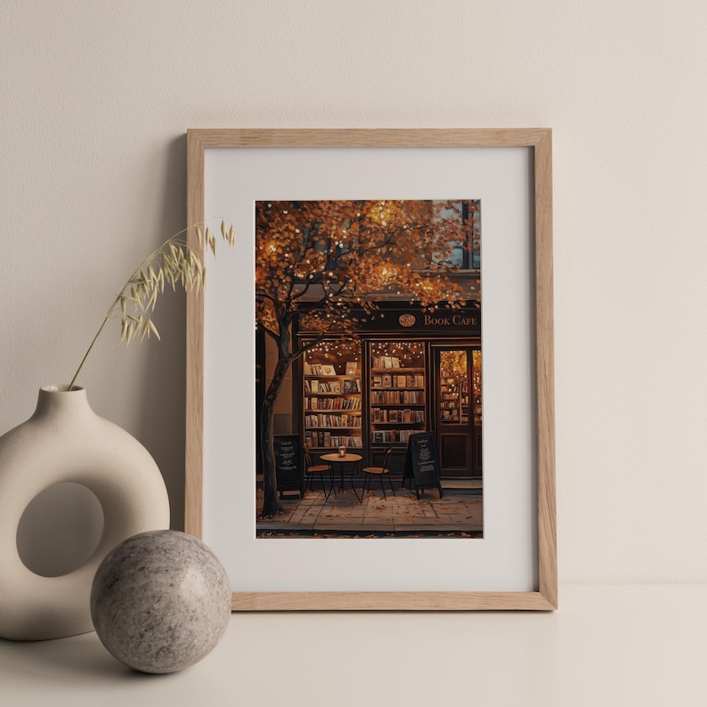 May include: A framed print of a cozy book cafe scene. The image features a storefront with the words "Book Cafe" above the entrance, bookshelves, a small table, and chairs. The scene is illuminated by warm lighting and surrounded by a tree with autumn leaves.