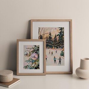 May include: Two framed art prints in light wood frames. The smaller print shows two disco ball ornaments hanging on a pine tree. The larger print depicts a winter scene with people ice skating on a rink surrounded by trees with string lights.