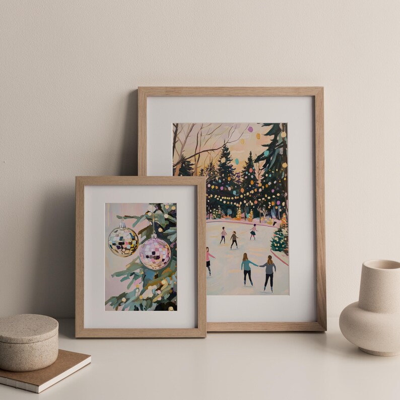May include: Two framed art prints in light wood frames. The smaller print shows two disco ball ornaments hanging on a pine tree. The larger print depicts a winter scene with people ice skating on a rink surrounded by trees with string lights.