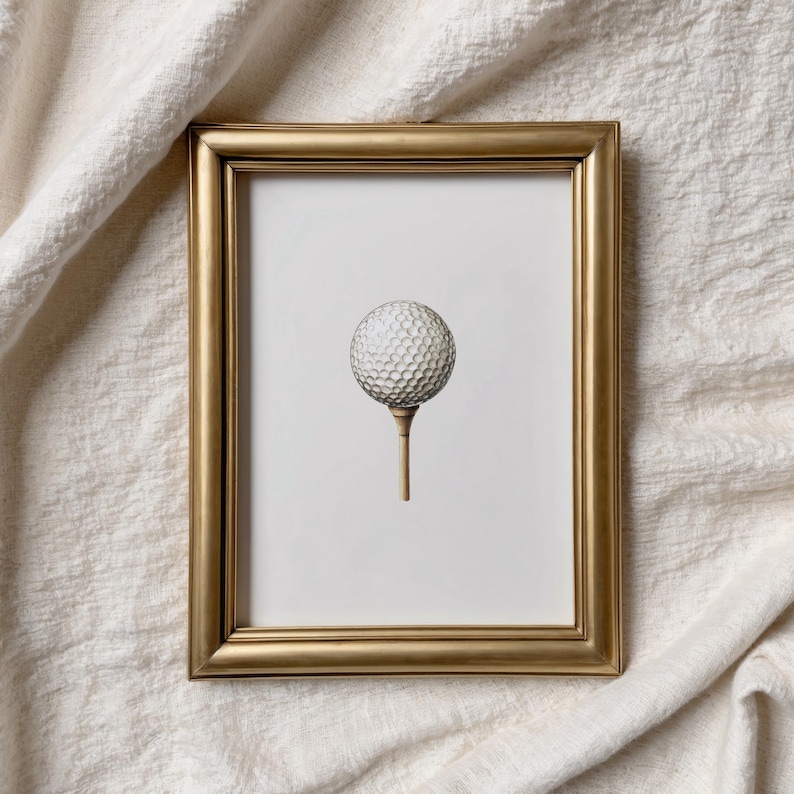 May include: A framed print featuring a golf ball on a tee. The golf ball is white with a dimpled surface, and the tee is a light brown color. The frame is gold, and the background is white.