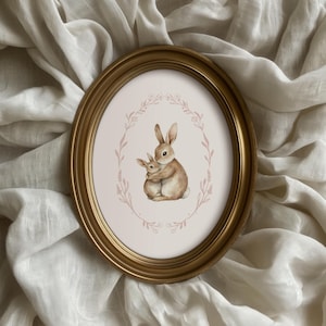 May include: An oval, gold-framed print featuring a watercolour illustration of two brown bunnies embracing. The bunnies are surrounded by a pink floral wreath on a white background. The frame is set against a backdrop of white fabric.