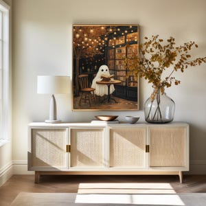 May include: A framed artwork depicts a ghost reading at a cafe table, with a warm, inviting atmosphere. The scene is illuminated by string lights and a cozy interior. The artwork hangs above a white cabinet with woven door panels, complemented by a table lamp and decorative vase.