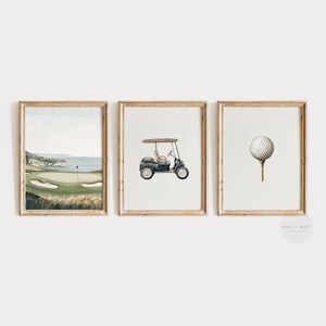 May include: Set of three framed watercolor prints with a golf theme. The prints feature a golf course landscape, a golf cart, and a golf ball on a tee. The frames are a light brown color.