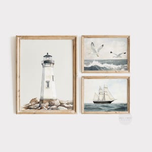 May include: Set of three framed watercolor prints featuring nautical themes. Includes a lighthouse on rocks, seagulls over ocean waves, and a sailboat on the water. The frames are light brown.