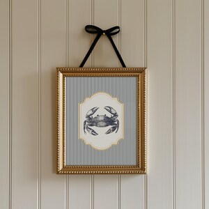 May include: A framed print with a nautical theme, depicting a crab illustration. The crab is on a cream-coloured, ornate shape, set against a blue and white striped background. The gold frame is suspended by a black ribbon.