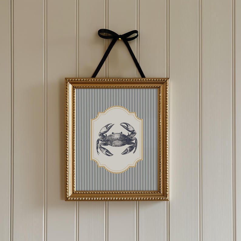 May include: A framed print with a nautical theme, depicting a crab illustration. The crab is on a cream-coloured, ornate shape, set against a blue and white striped background. The gold frame is suspended by a black ribbon.
