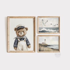 May include: A set of three framed wall art prints featuring a brown teddy bear wearing a navy blue shirt and white overalls with a sailor hat, a seascape with two white seagulls flying over ocean waves, and a lighthouse on a grassy coastal cliff.
