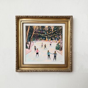 May include: A framed painting depicting a group of people ice skating on an outdoor rink surrounded by trees decorated with glowing string lights. The artwork is enclosed in an ornate, metallic gold frame with a white mat border.