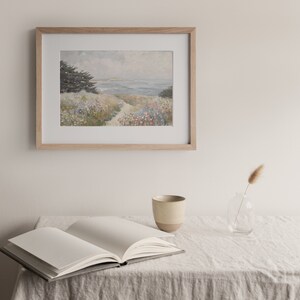 May include: A framed landscape painting depicts a coastal scene with a path leading to the ocean. The artwork is in a light wood frame, hanging above an open book, a ceramic mug, and a small glass vase with a dried plant on a linen tablecloth.