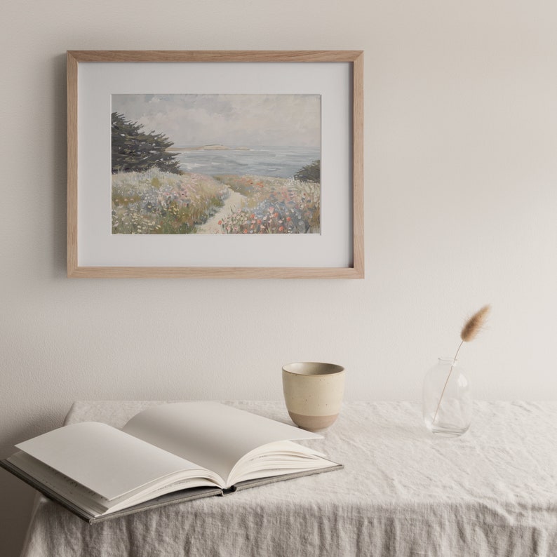 May include: A framed landscape painting depicts a coastal scene with a path leading to the ocean. The artwork is in a light wood frame, hanging above an open book, a ceramic mug, and a small glass vase with a dried plant on a linen tablecloth.