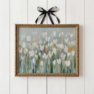 Puede incluir: Un cuadro rectangular con marco dorado de cuentas que muestra un campo de tulipanes blancos, crema y naranja suave sobre un fondo verde salvia. Una cinta de sat&eacute;n negro est&aacute; atada en un lazo en la parte superior central para colgarlo.